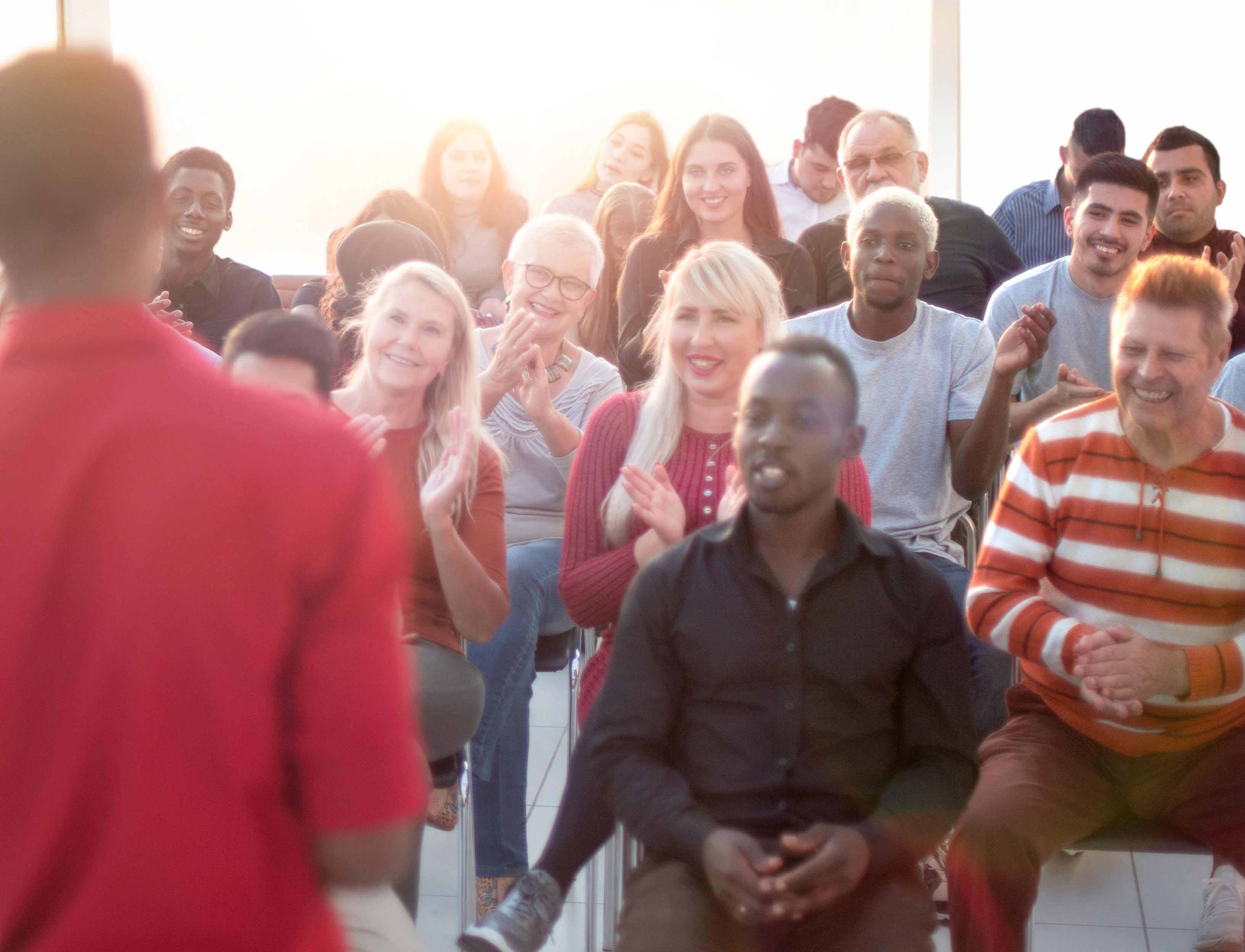 Audience of diverse people smiling and clapping while listening to a speaker in red shirt.