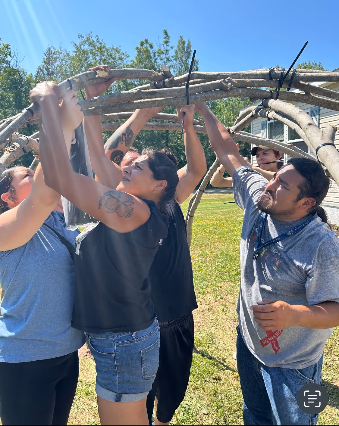 Group of people outdoors assembling a dome structure with wooden branches under a clear blue sky.