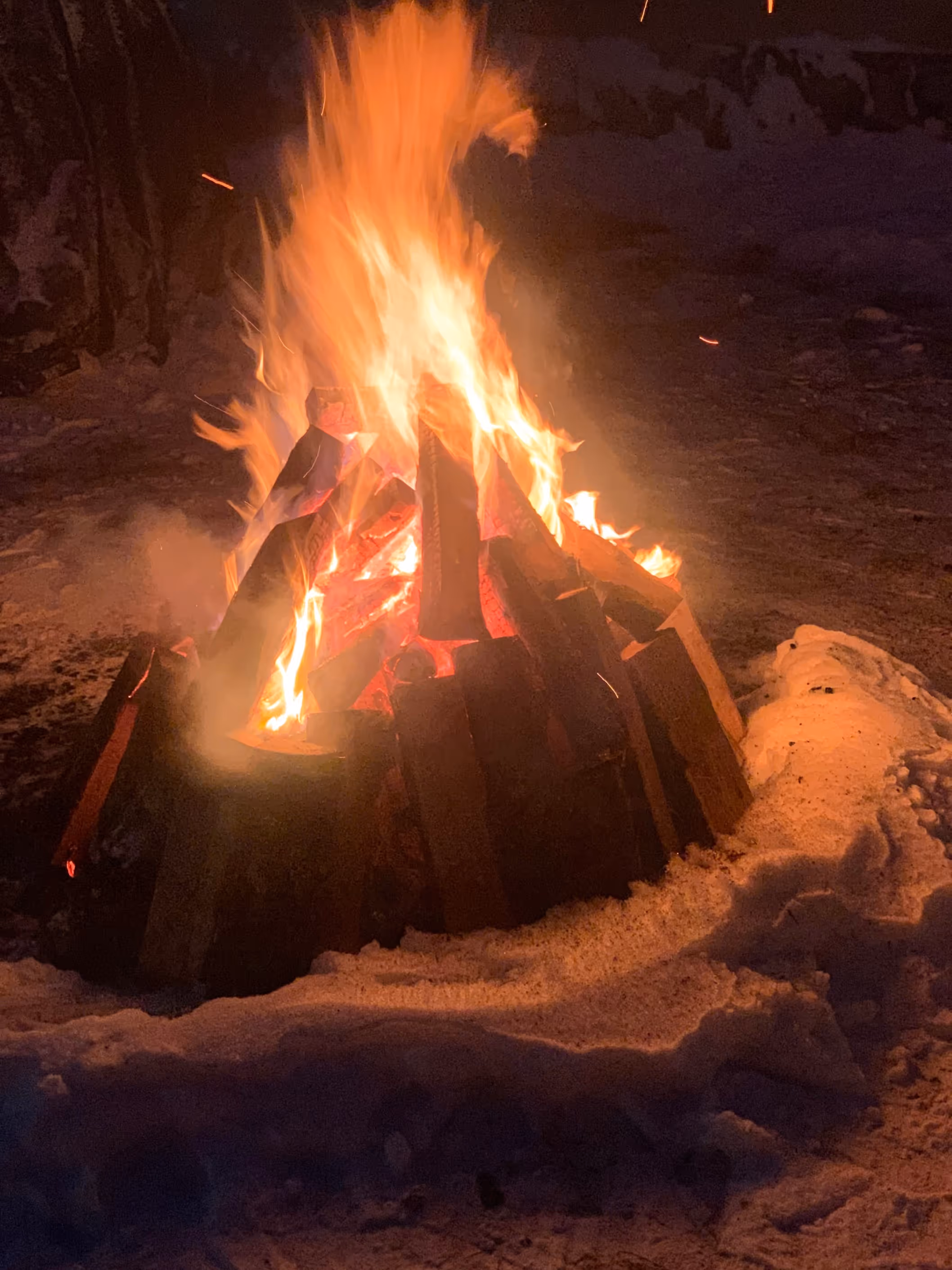 A bright campfire burning wooden logs surrounded by snow at night.