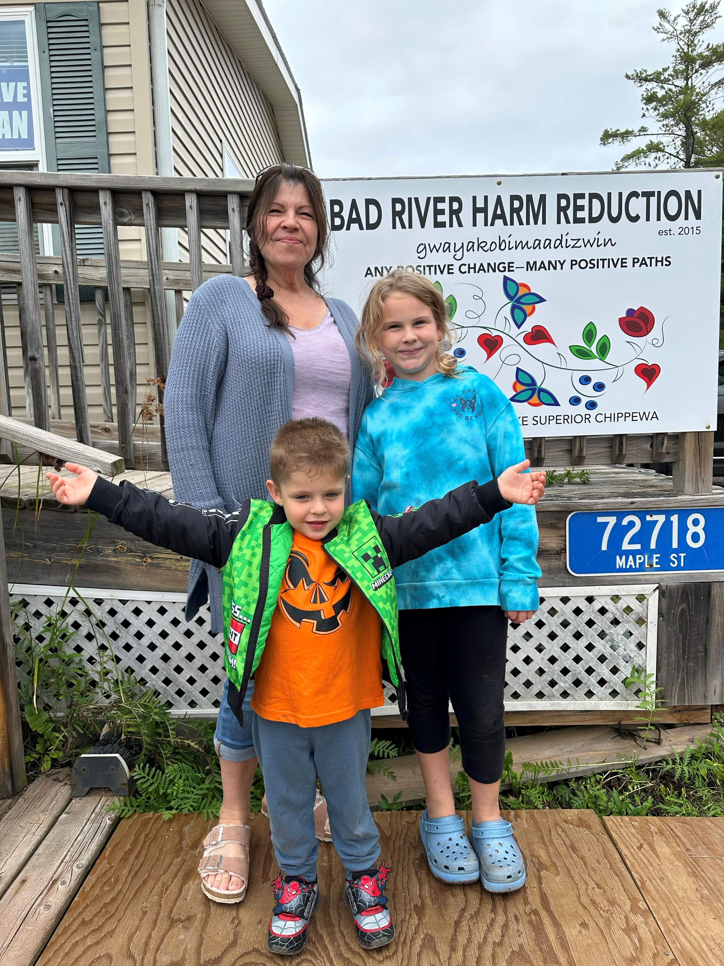 Woman with two children standing on wooden porch in front of Bad River Harm Reduction sign.