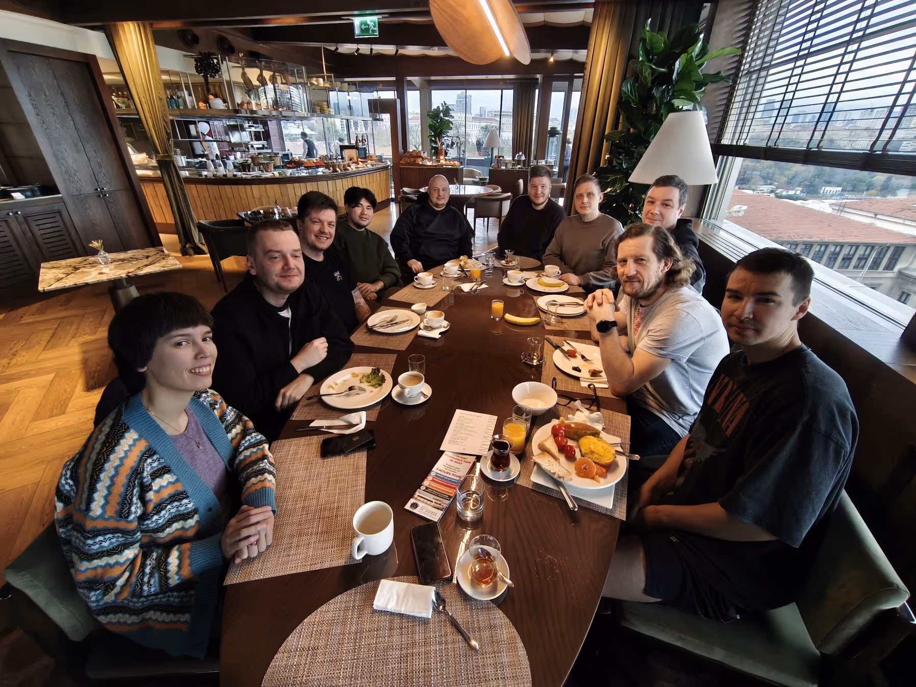 Group of ten people sitting around a dining table with plates, drinks, and city view outside windows.