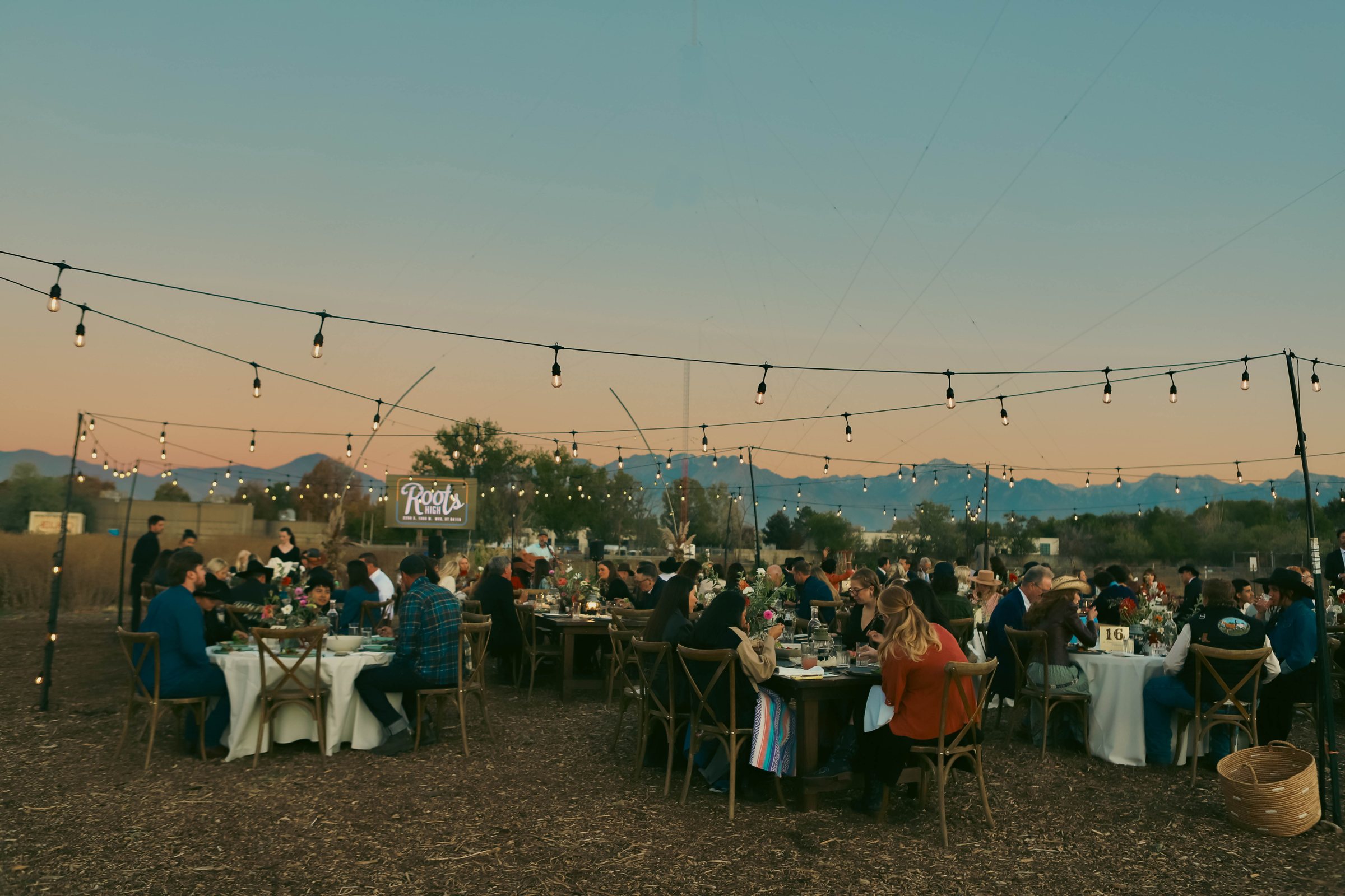 Guests at long tables in the Roots flower field at dusk, string lights strung overhead and the Wasatch range behind them.