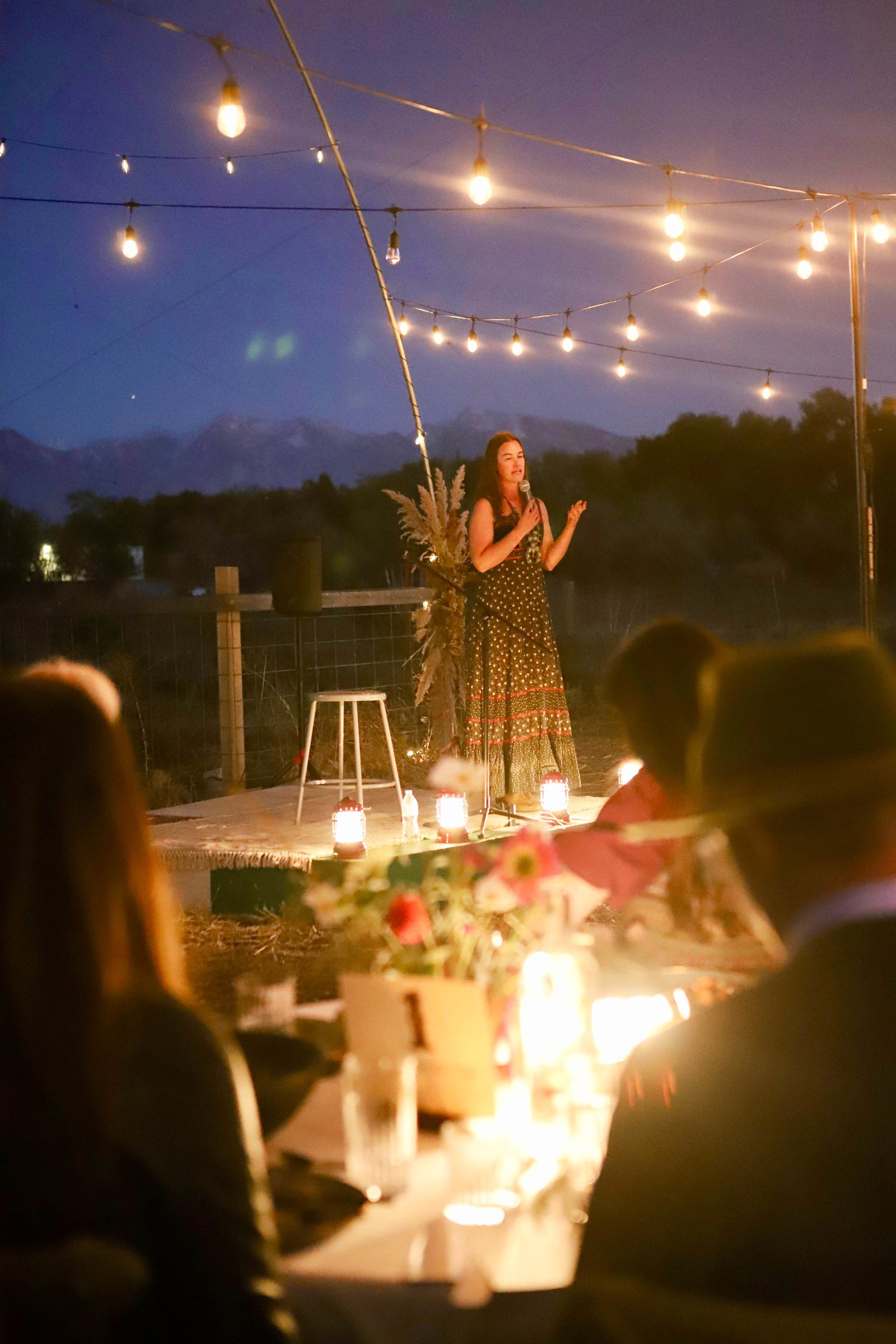A speaker at the microphone on a low stage at blue hour, lanterns at her feet and guests listening from candlelit tables.