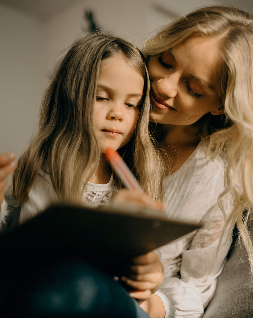 Blonde woman smiling and looking at a young girl who is writing on a clipboard with a red pen.