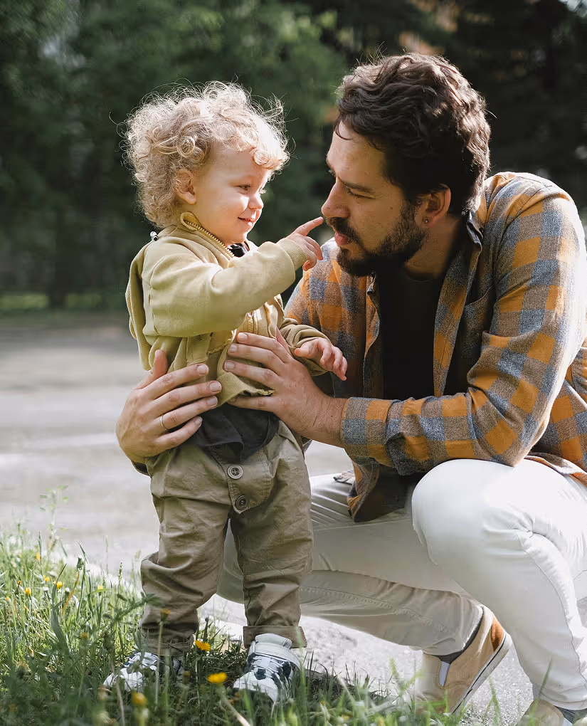 Toddler in khaki pants and yellow hoodie touching nose of a man with beard wearing a plaid jacket in an outdoor setting.