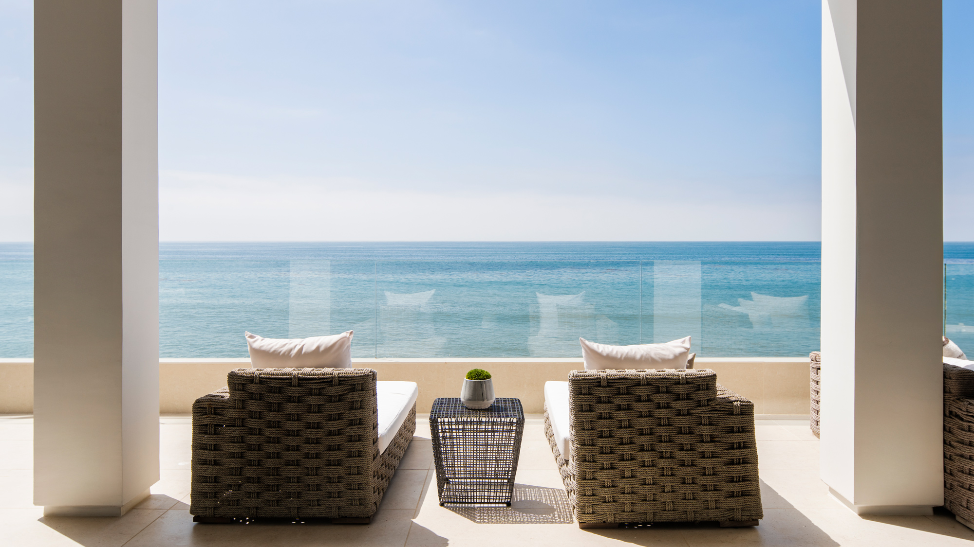Two wicker lounge chairs with white cushions face ocean on a sunny patio with glass railing.