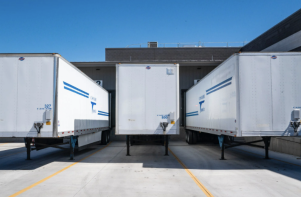 Three white semi-truck trailers parked at loading docks under a clear blue sky.