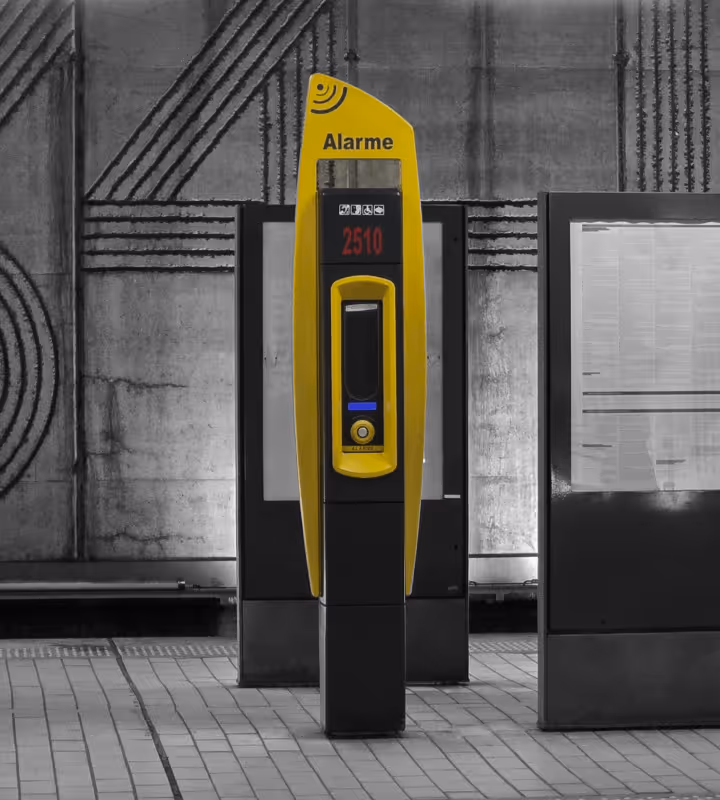 Borne d'alarme jaune sur un quai de métro avec un panneau d'affichage en arrière-plan.