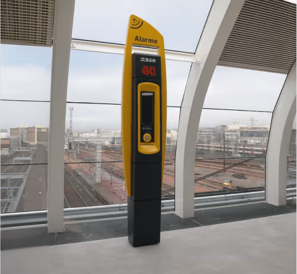 Poste d'alarme jaune et noir dans une gare avec rails visibles à l'extérieur.