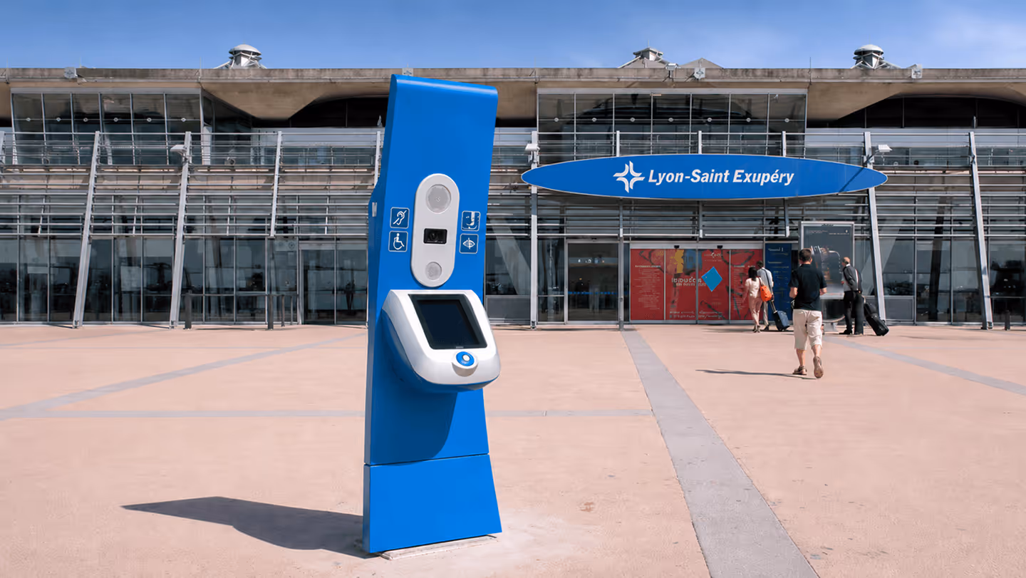 Borne d'information bleue accessible devant l'entrée de l'aéroport Lyon-Saint Exupéry avec des passagers marchant.