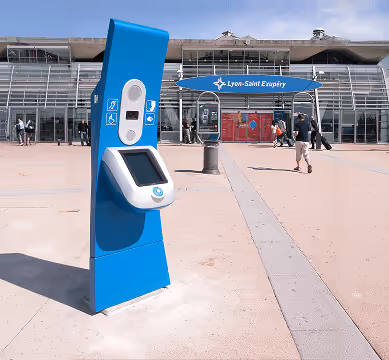 Borne interactive bleue devant l'entrée de l'aéroport Lyon-Saint Exupéry avec des passants.
