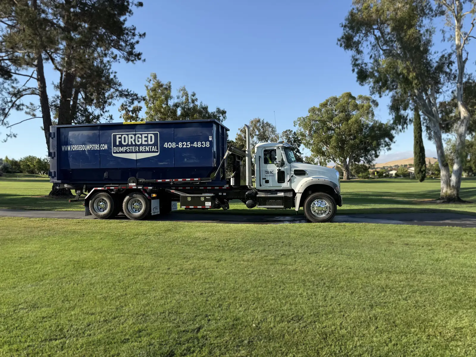 Forged Dumpster Rental roll-off truck hauling a navy dumpster across a grassy lot in San Jose