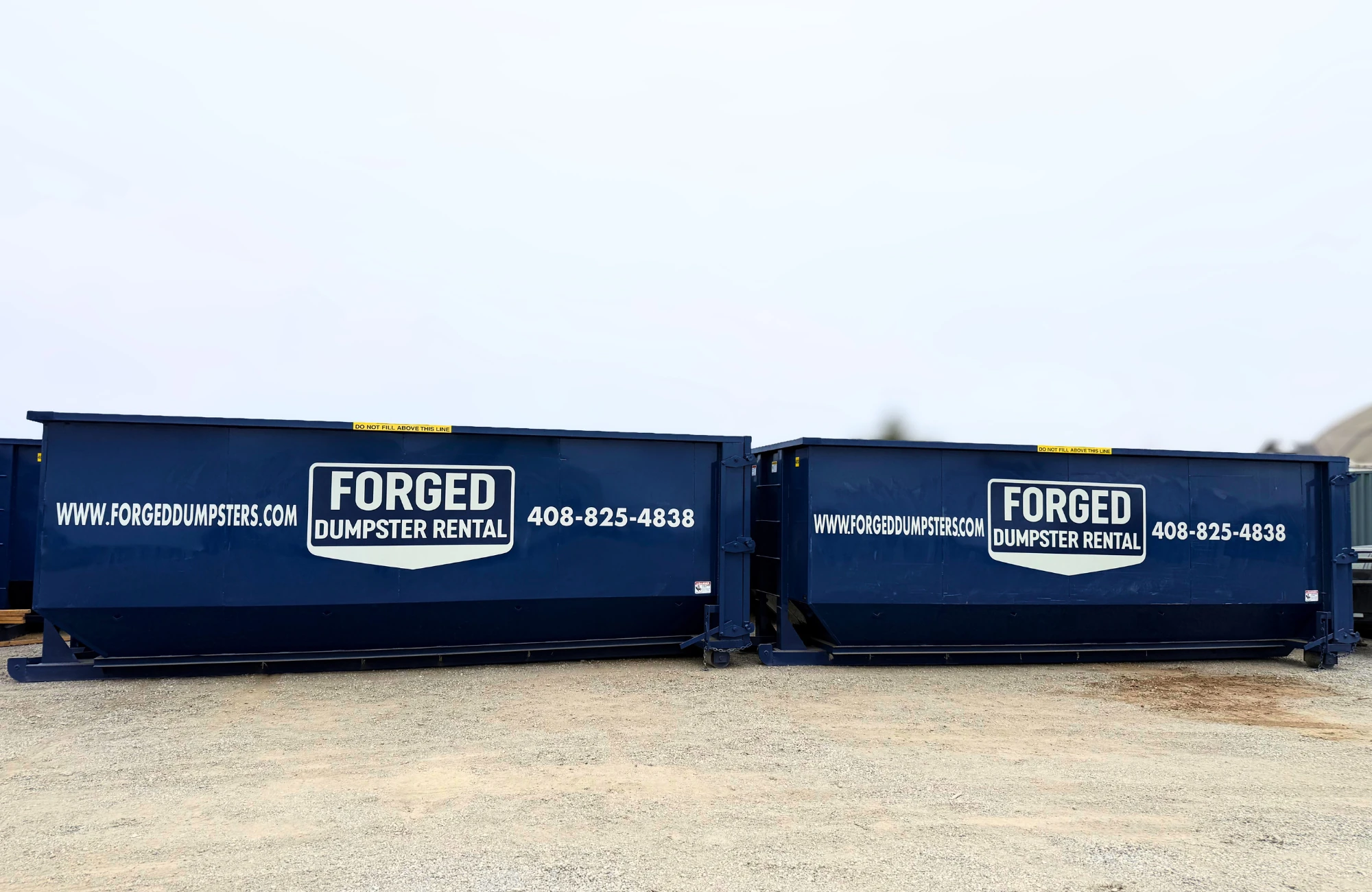 Two navy Forged Dumpster Rental roll-off containers parked on a gravel lot