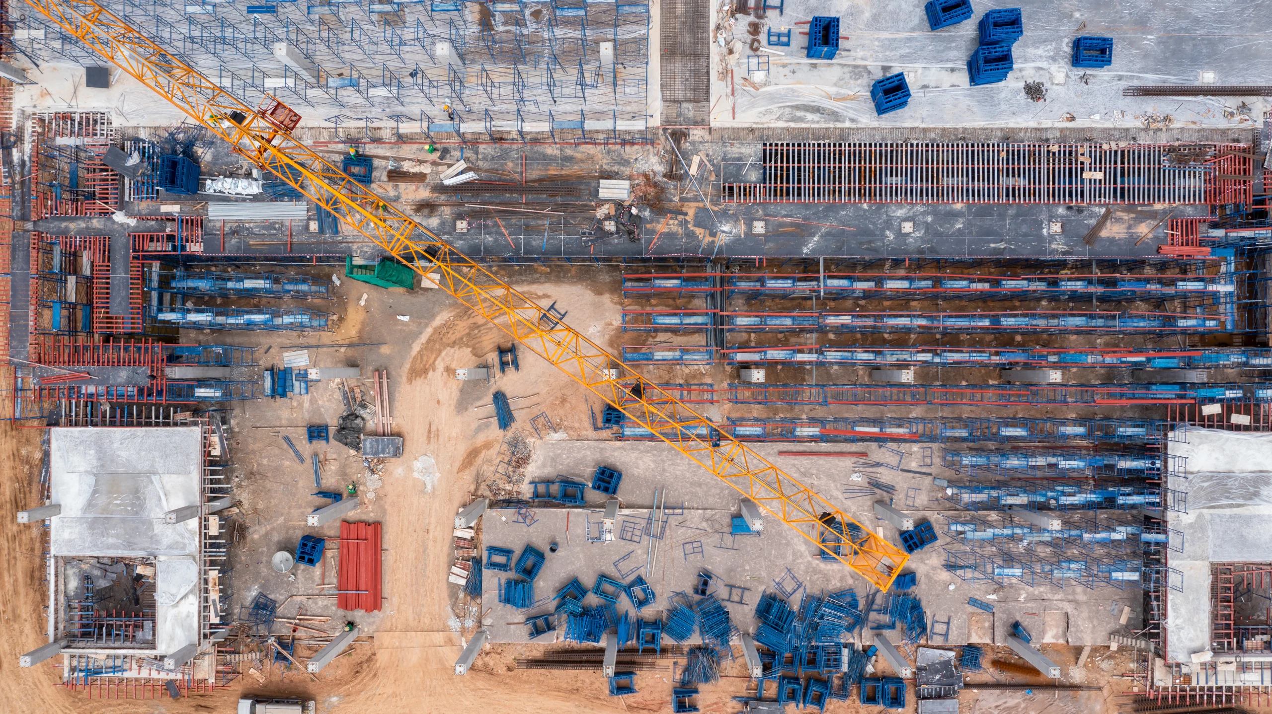 Aerial view of a large construction site with a yellow tower crane, rebar, formwork and stacked materials