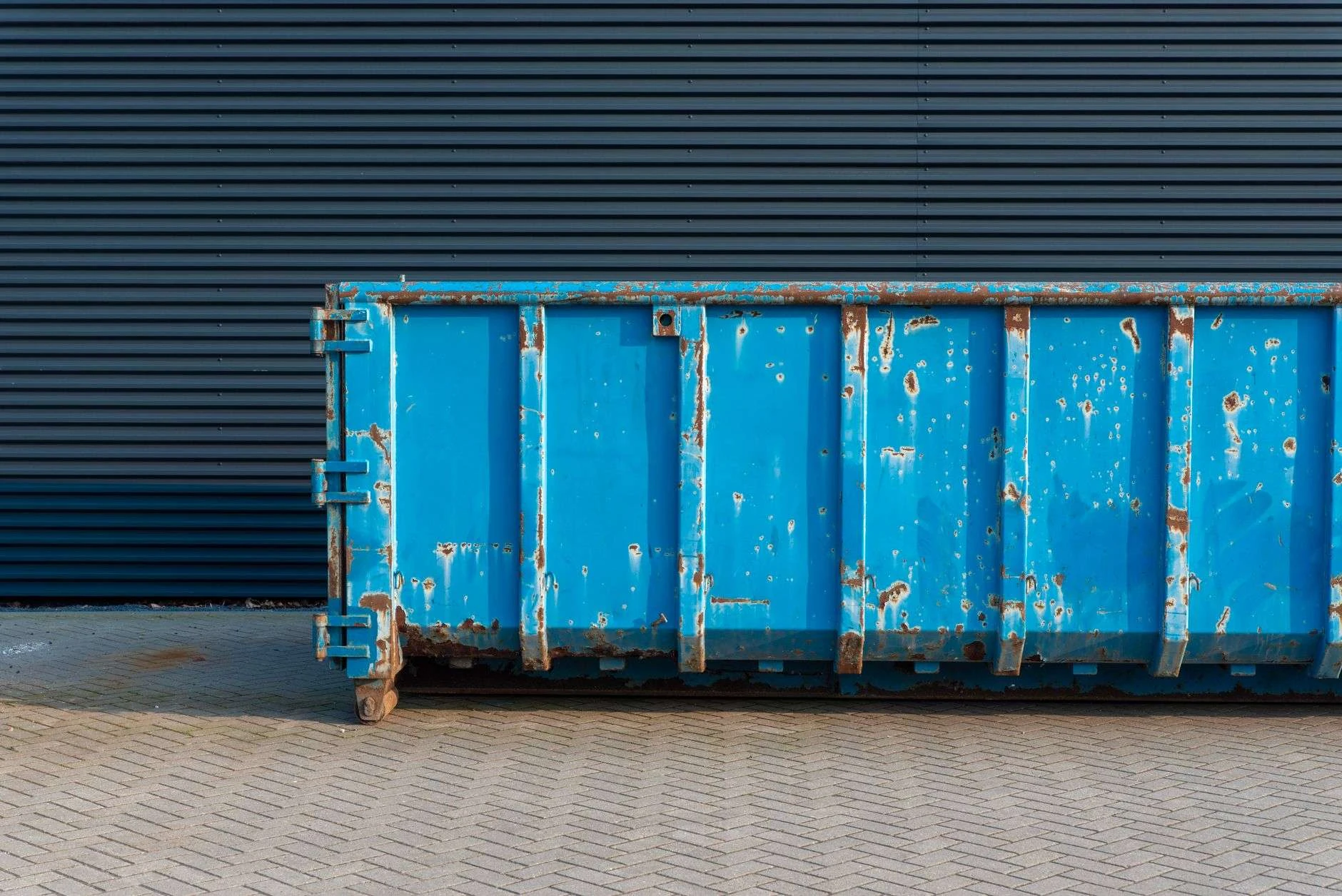 Weathered blue roll-off dumpster container parked on brick pavers against a dark corrugated metal wall