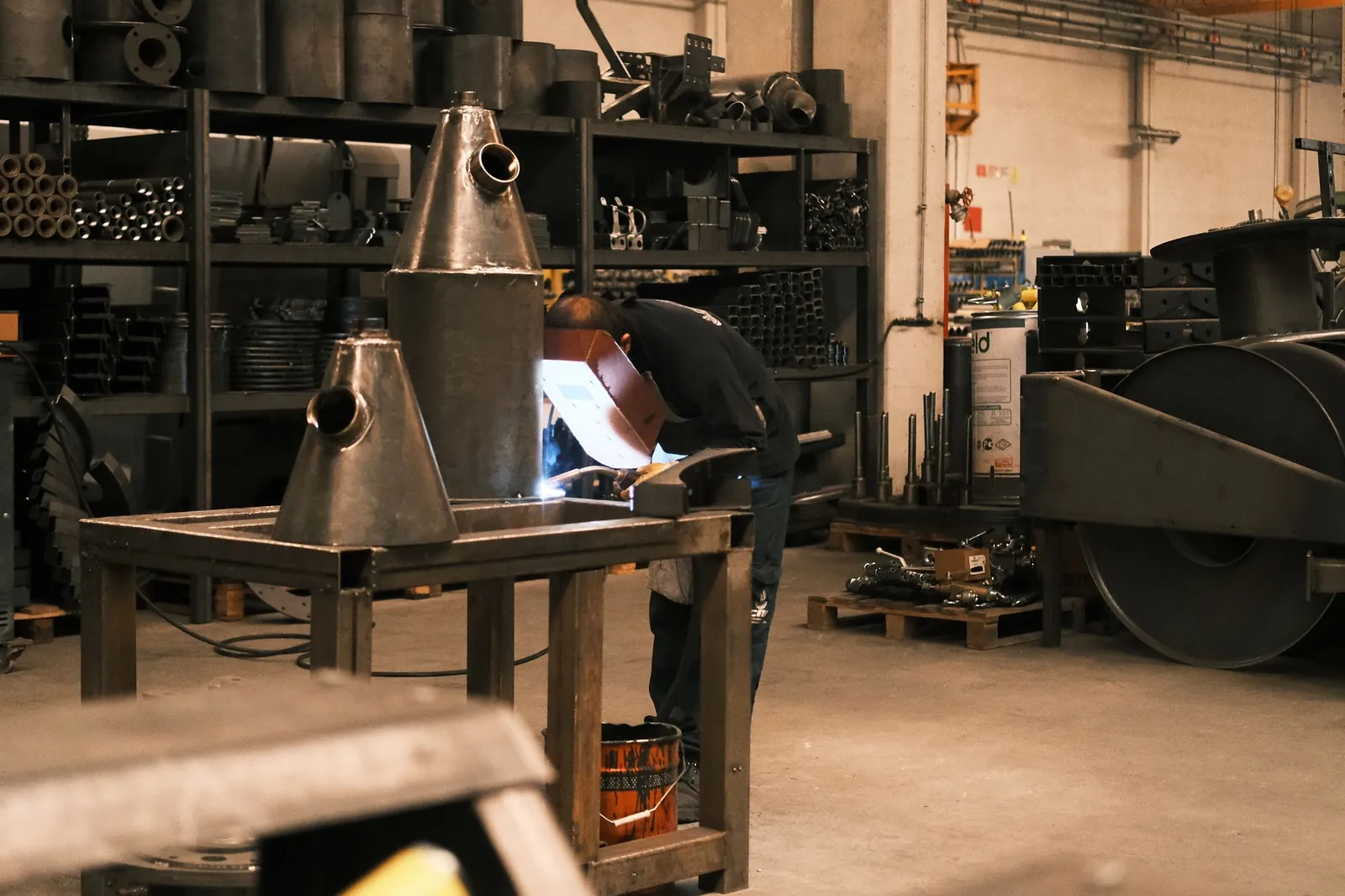 Welder joining sheet metal in a fabrication shop with racks of steel tube, plate and offcuts behind him