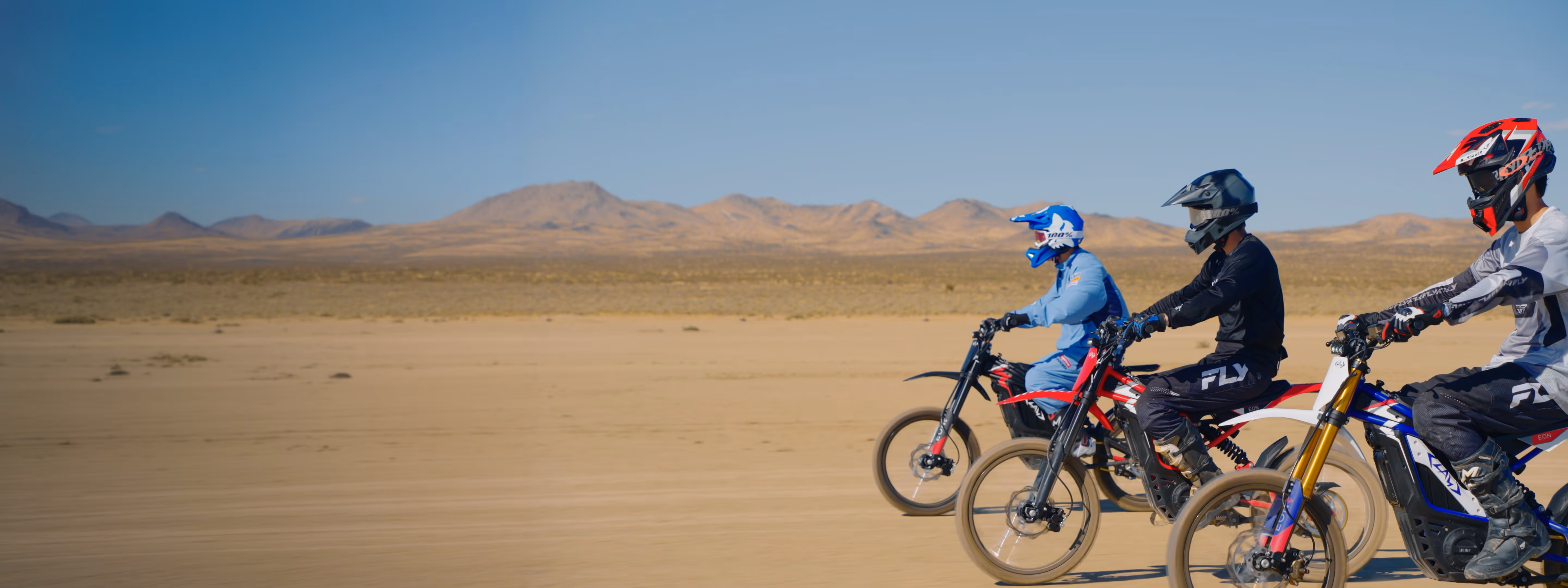 Three dirt bike riders wearing helmets riding side by side in a desert landscape with mountains.