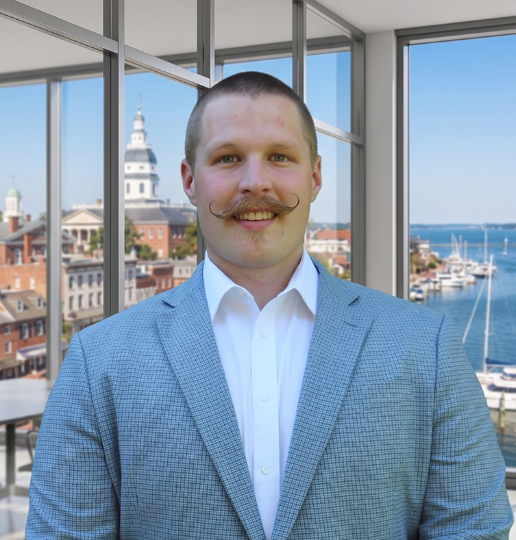 Peyen in a gray suit and white shirt standing in front of windows overlooking a harbor.