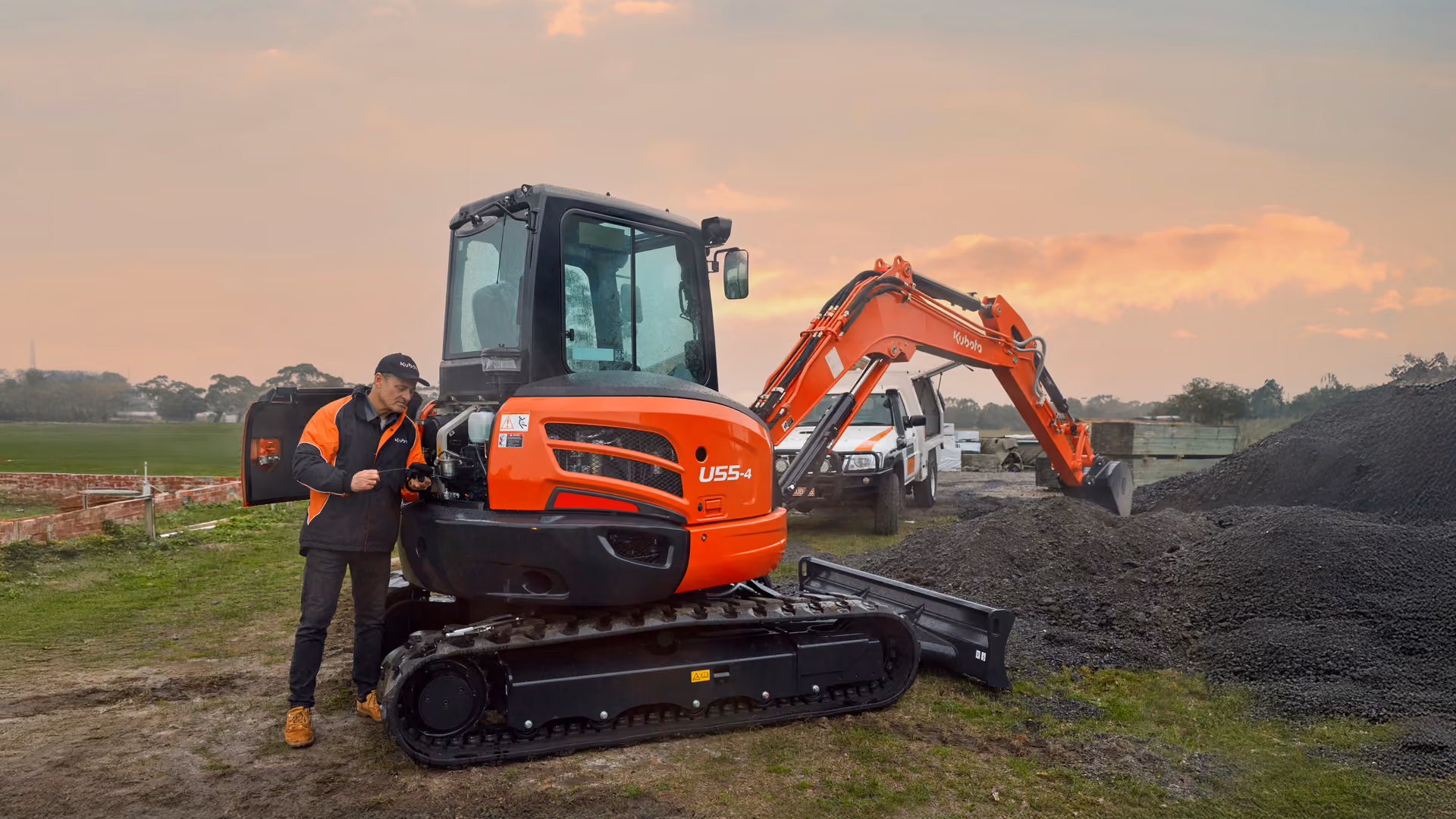 Kubota U55-4 excavator and operator on a work site at dusk, with a service vehicle in the background.
