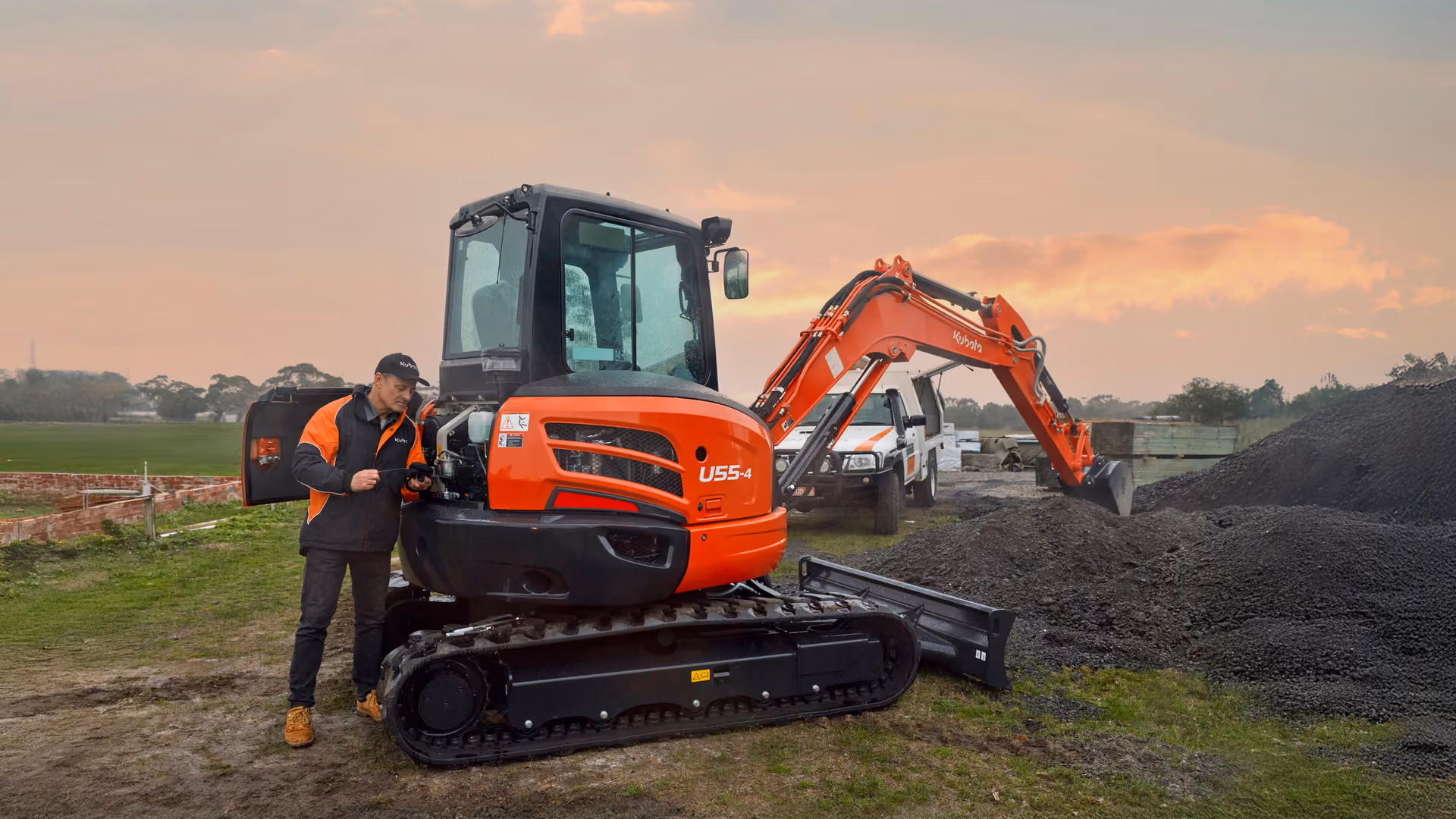 Operator beside a Kubota excavator on a work site at sunset