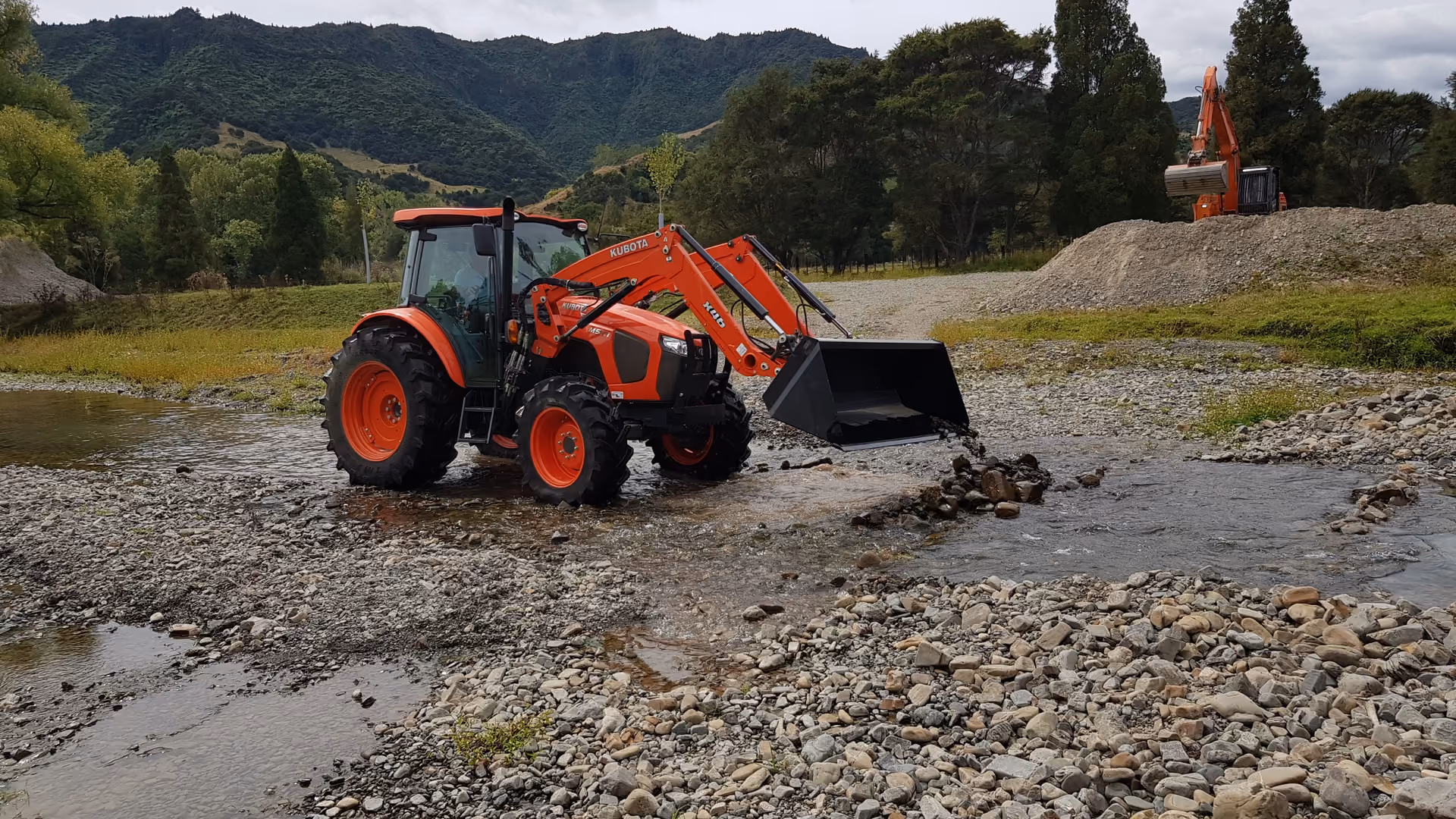Kubota tractor with front loader working a riverbed below bush-clad hills