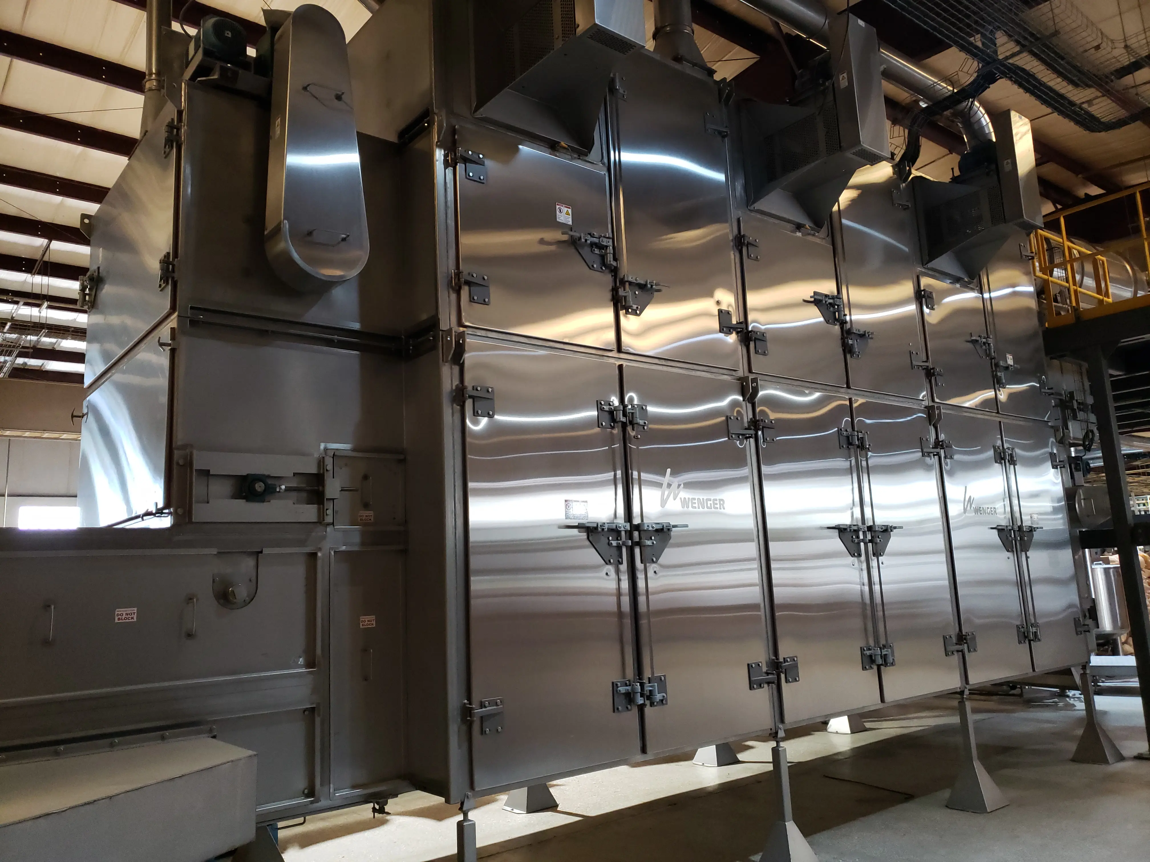 Large industrial stainless steel machine with multiple doors and ventilation fans inside a warehouse.