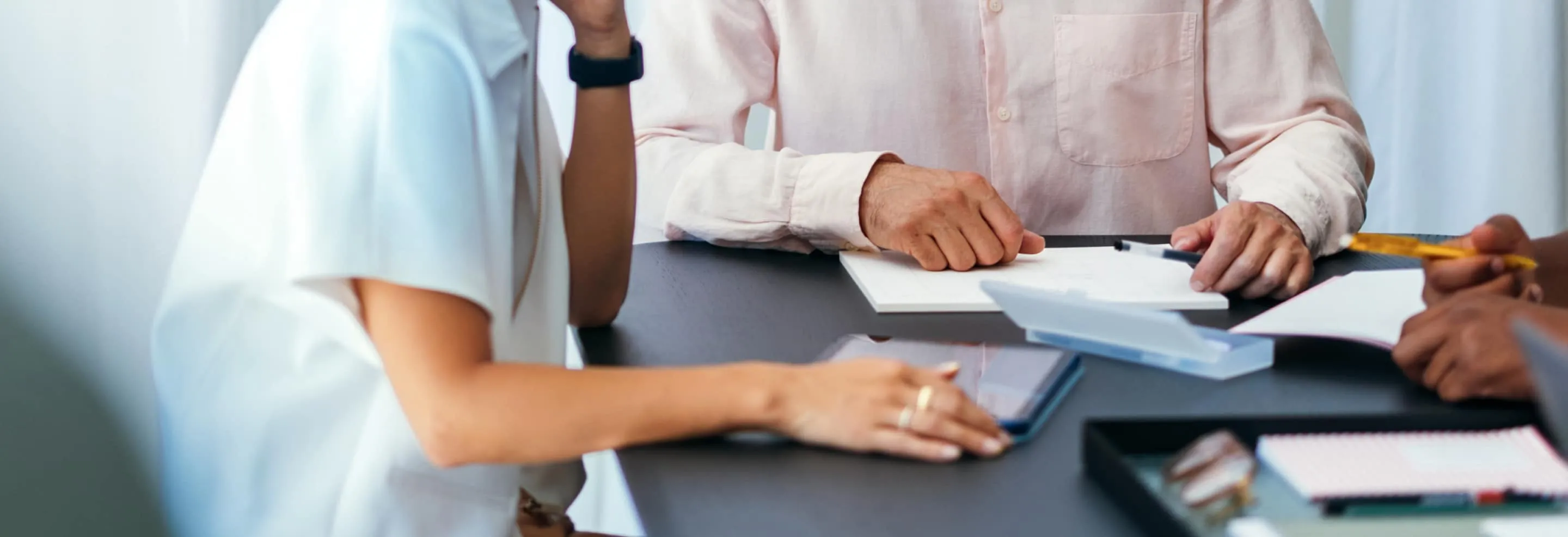 Three people at a table engaged in discussion with notepads, pens, and a smartphone visible.