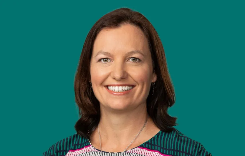 Smiling woman with brown hair wearing a colorful striped top and silver necklace on teal background.