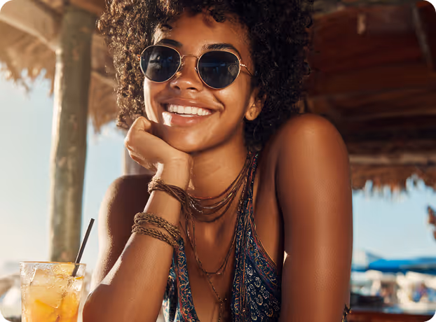 Smiling woman with curly hair and sunglasses sitting at a beach bar with a drink nearby.