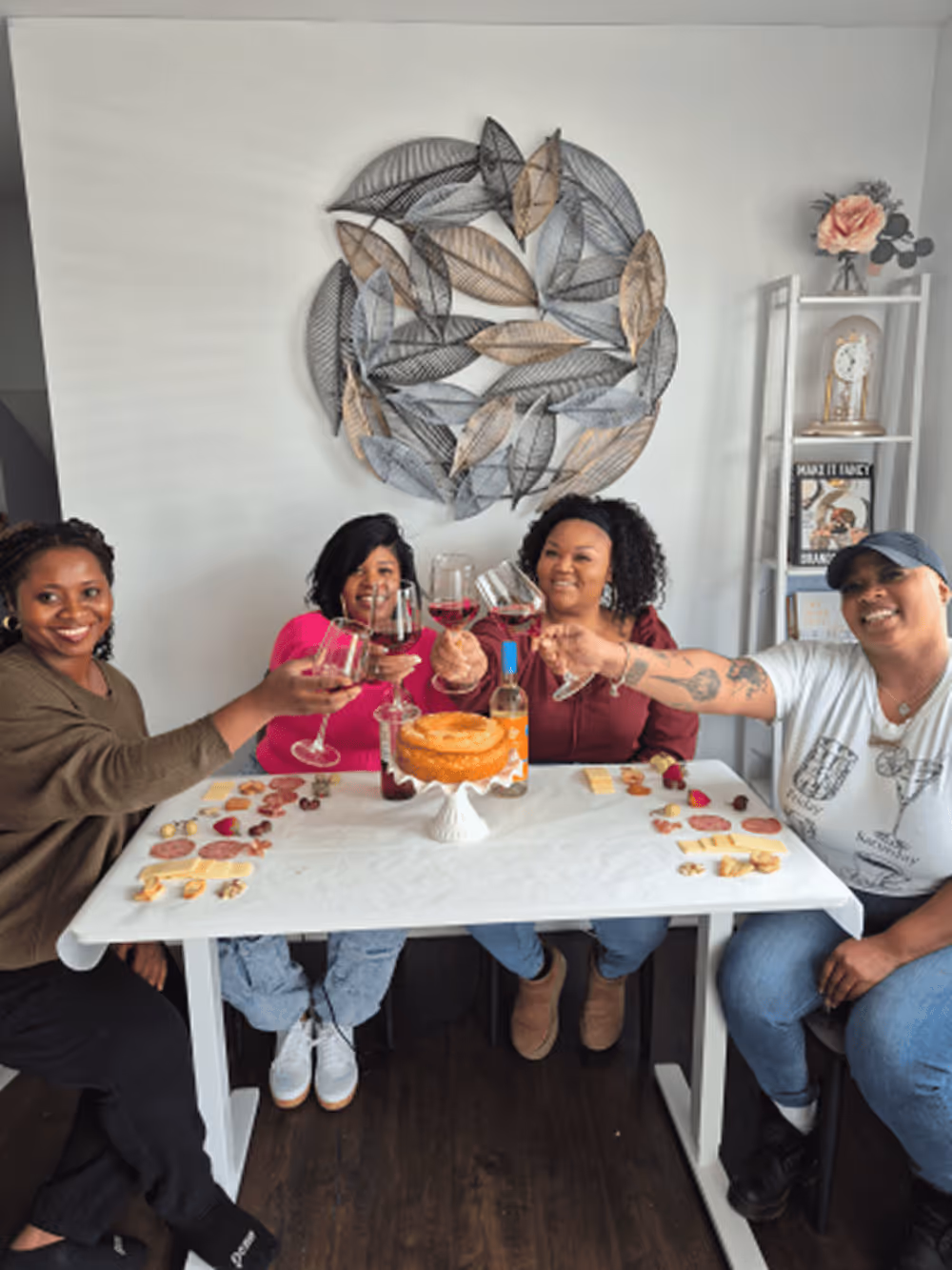 Four women sitting around a table raising glasses of red wine in a toast, with a cake, charcuterie, and wine bottle on the table.