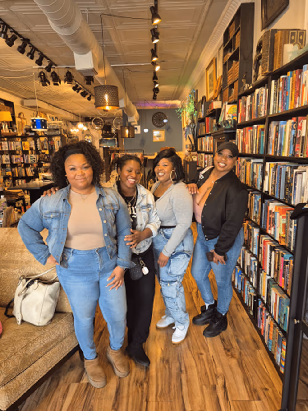 Four women smiling and posing together in a cozy bookstore with wooden floors and bookshelves.