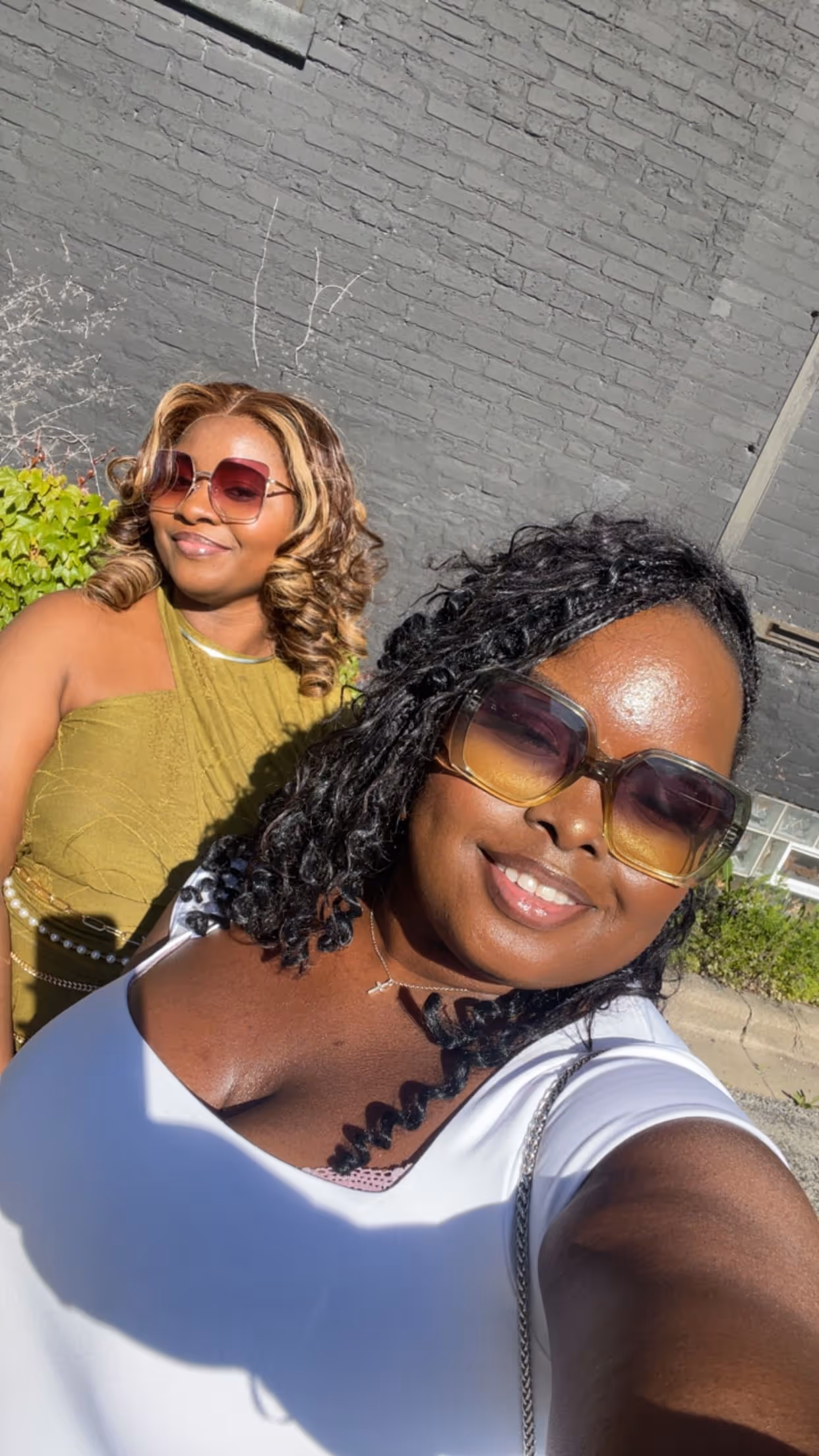 Two women smiling outdoors, both wearing sunglasses; one in a white top with curly hair and the other in a green halter dress with wavy highlights.