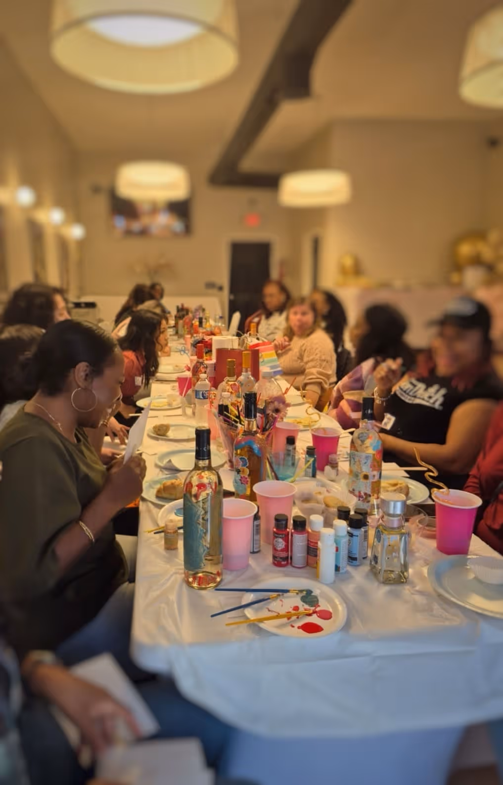 Group of people seated at a table painting and decorating wine bottles during a craft workshop.