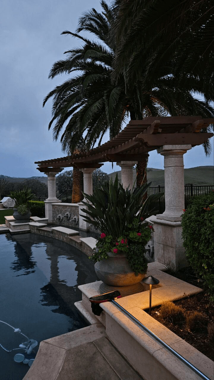 Elegant pergola in San Jose featuring sturdy stone columns and a wooden slatted roof, overlooking a glowing pool.