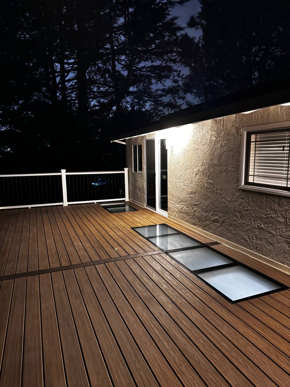 Stunning nighttime deck with warm ambient lighting, glowing in-floor LED light panels, white railings, and a cozy house exterior. A luxurious outdoor living space perfect for evening relaxation and entertaining under the stars