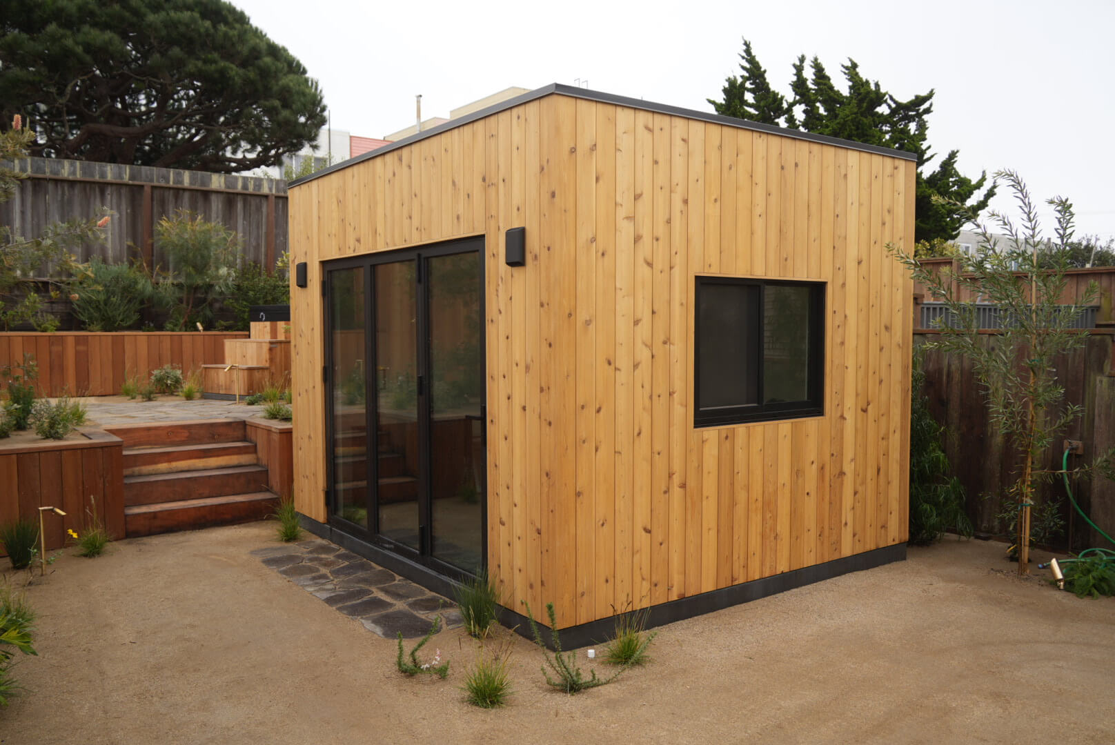 Modern cedar backyard shed in California with vertical wood siding, black-framed sliding glass doors, a matching side window, and minimalist black exterior lighting, set in a landscaped backyard.