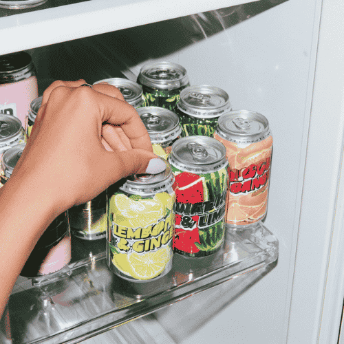 Hand grabbing a lemon ginger soda can from a fridge shelf with various flavored soda cans.