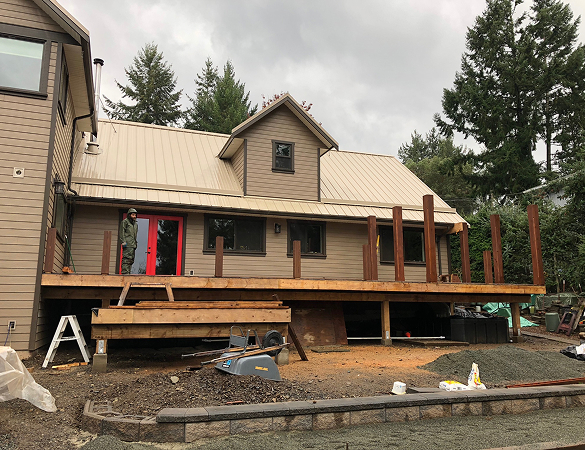 Construction of a wooden deck being built outside a beige house with a metal roof under cloudy sky.