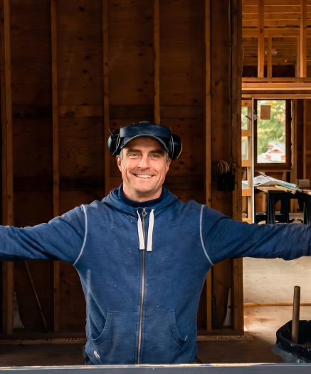 Smiling man in a black hoodie and cap with ear protection inside a wooden construction site.