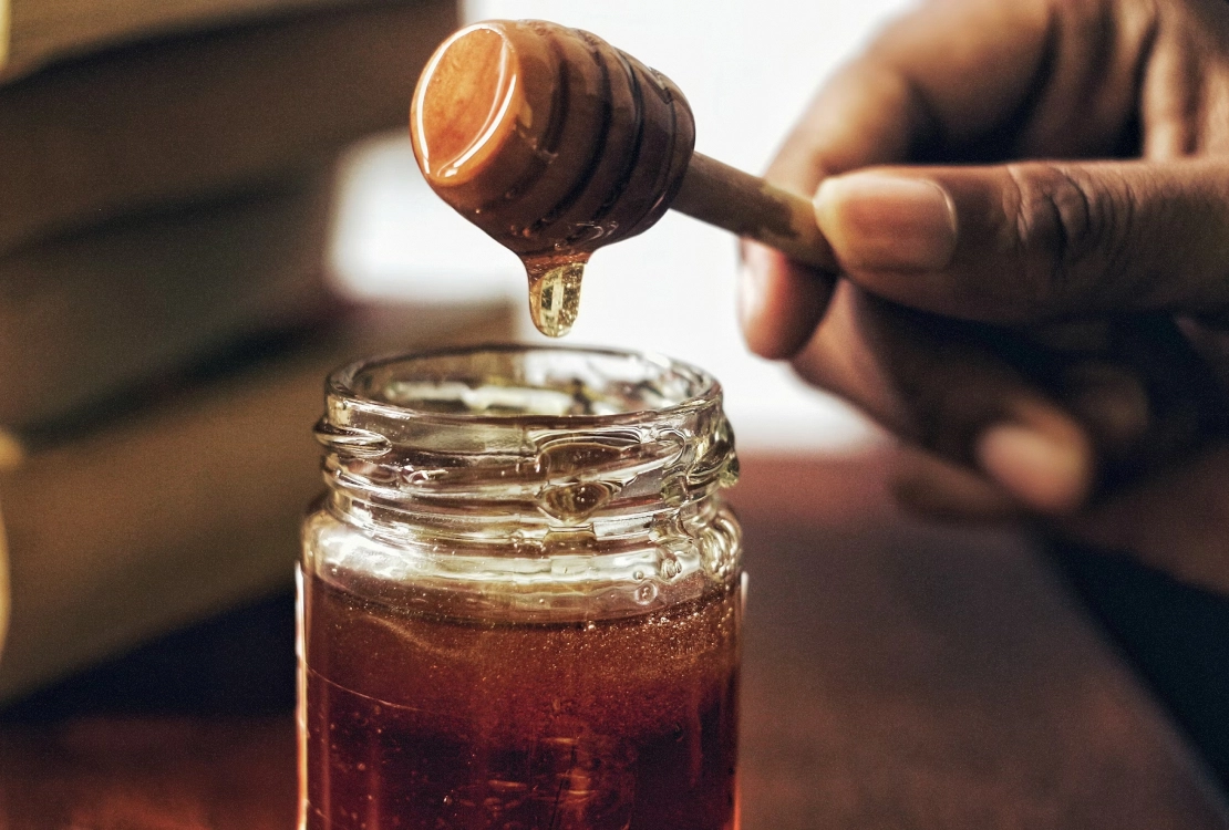 Hand holding honey dipper with honey dripping into a glass jar of honey.