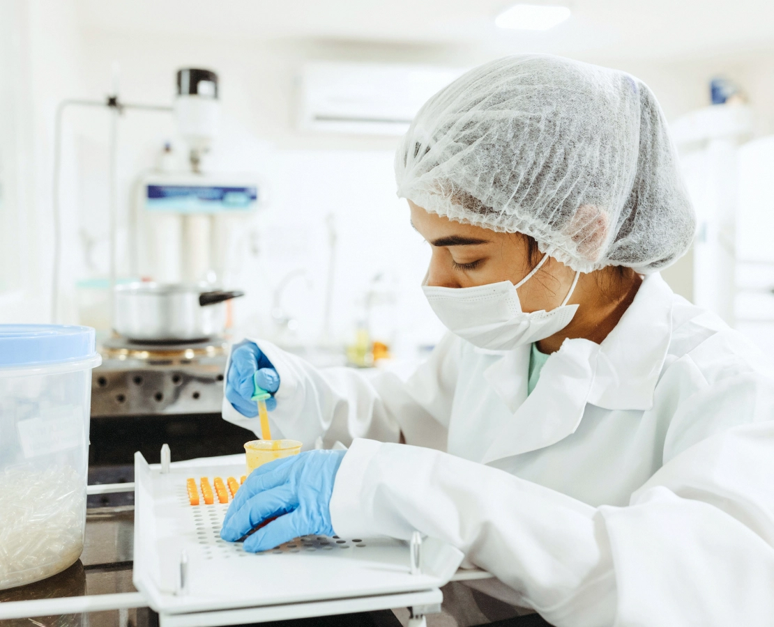 Scientist in lab coat, hairnet, and mask working with orange samples and yellow liquid in a laboratory.