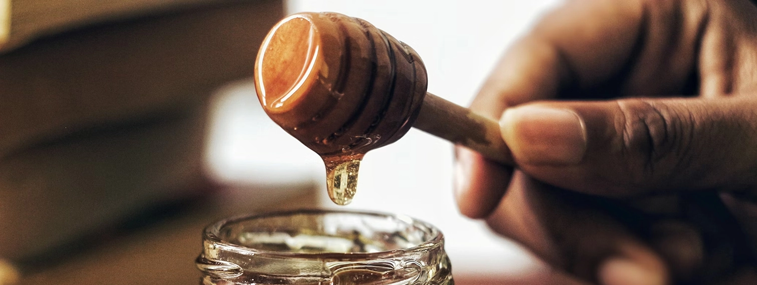 Hand holding wooden honey dipper with honey dripping over an open glass jar.