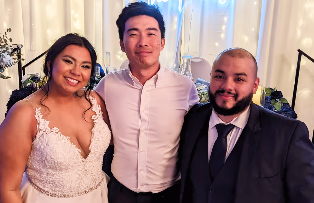 Bride in white dress, a man in white shirt, and a man in dark suit smiling and posing indoors.