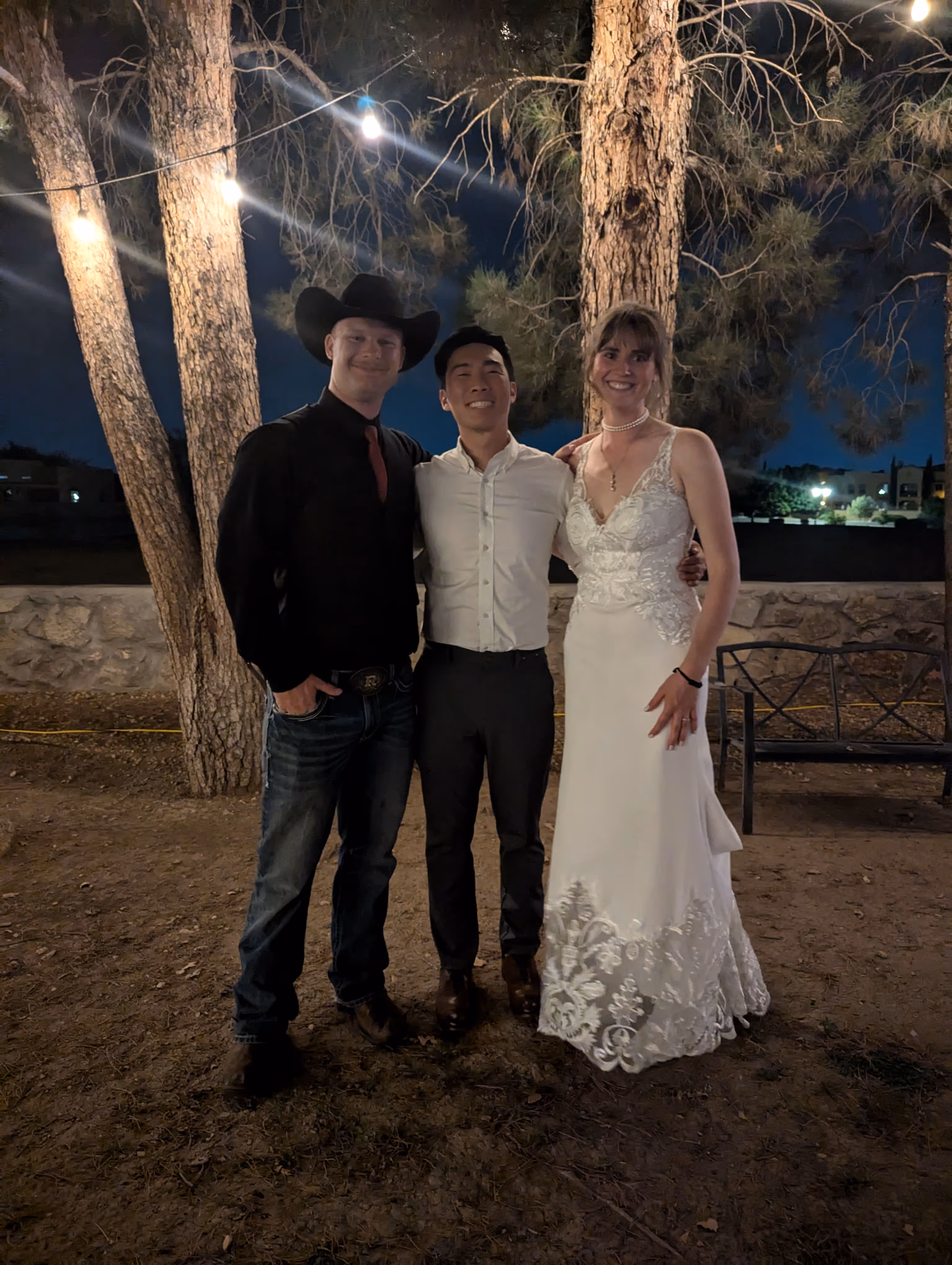 Three people smiling outdoors at night: a man in a cowboy hat, a man in a white shirt, and a woman in a white dress.