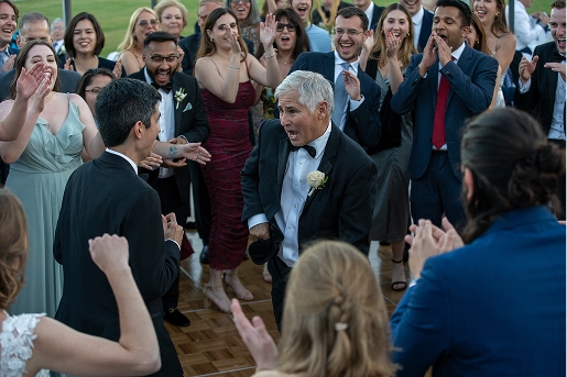 Group of well-dressed people clapping and cheering around two men dancing at an outdoor event.