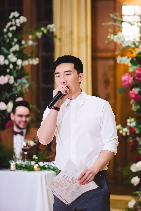 Man in white shirt speaking into a microphone while holding papers at an indoor event with floral decor.