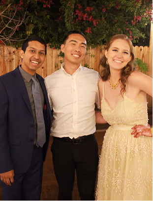 Three people smiling: two men dressed formally and a woman in a yellow lace dress with henna on her arm.