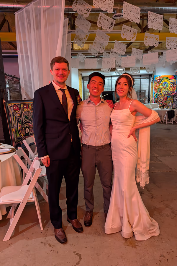 Three people posing indoors at a wedding: a groom, bride in white dress, and a man in a light shirt.