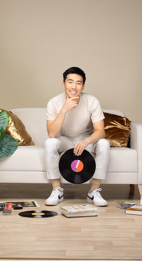 Smiling man sitting on sofa holding a vinyl record with more records and books on floor.