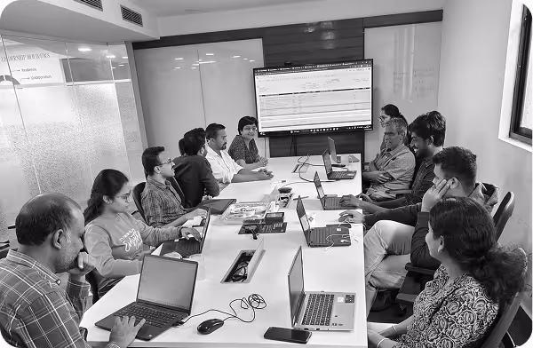 Group of people in a meeting room working on laptops with a screen displaying data at the front.